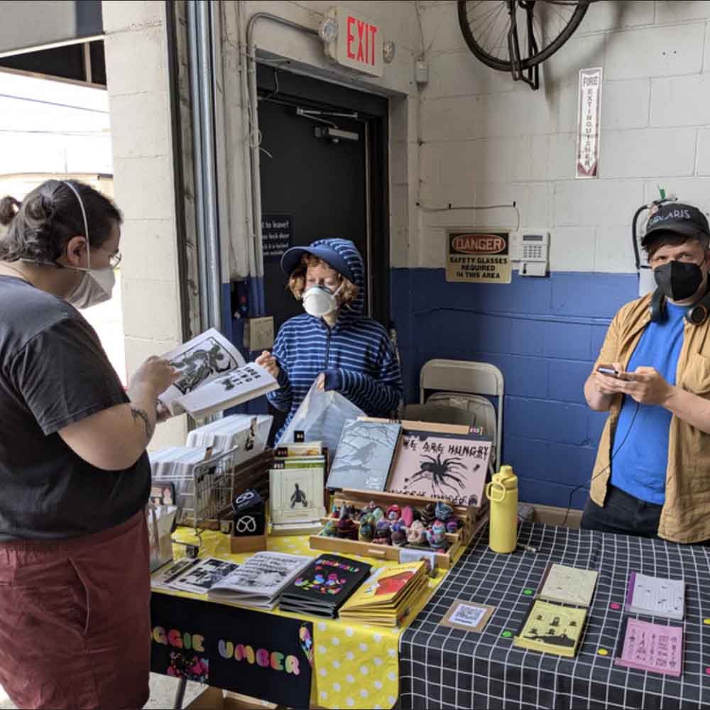 Maggie Umber tabling at Insert Name Zine Fest 2024 talking to Ezra David Mattes, standing next to Justin Skarhus of Entropy Editions, photo by Ryan C